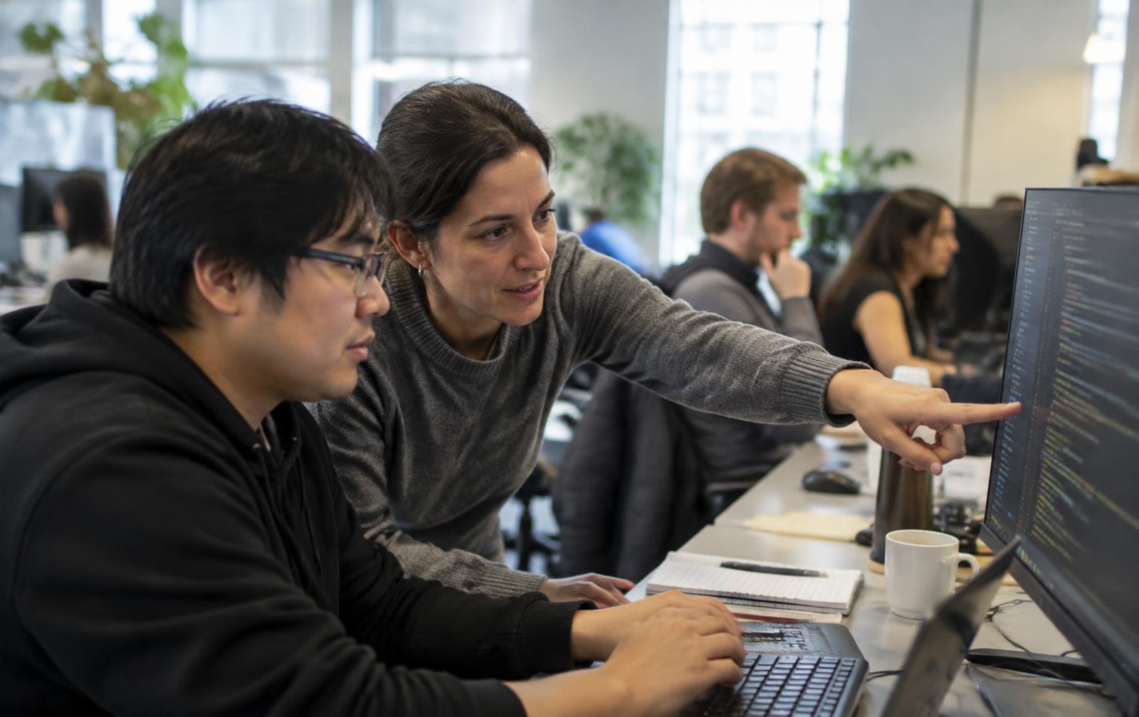 Engineering manager leaning in to review code on a monitor with a software engineer in an open engineering workspace