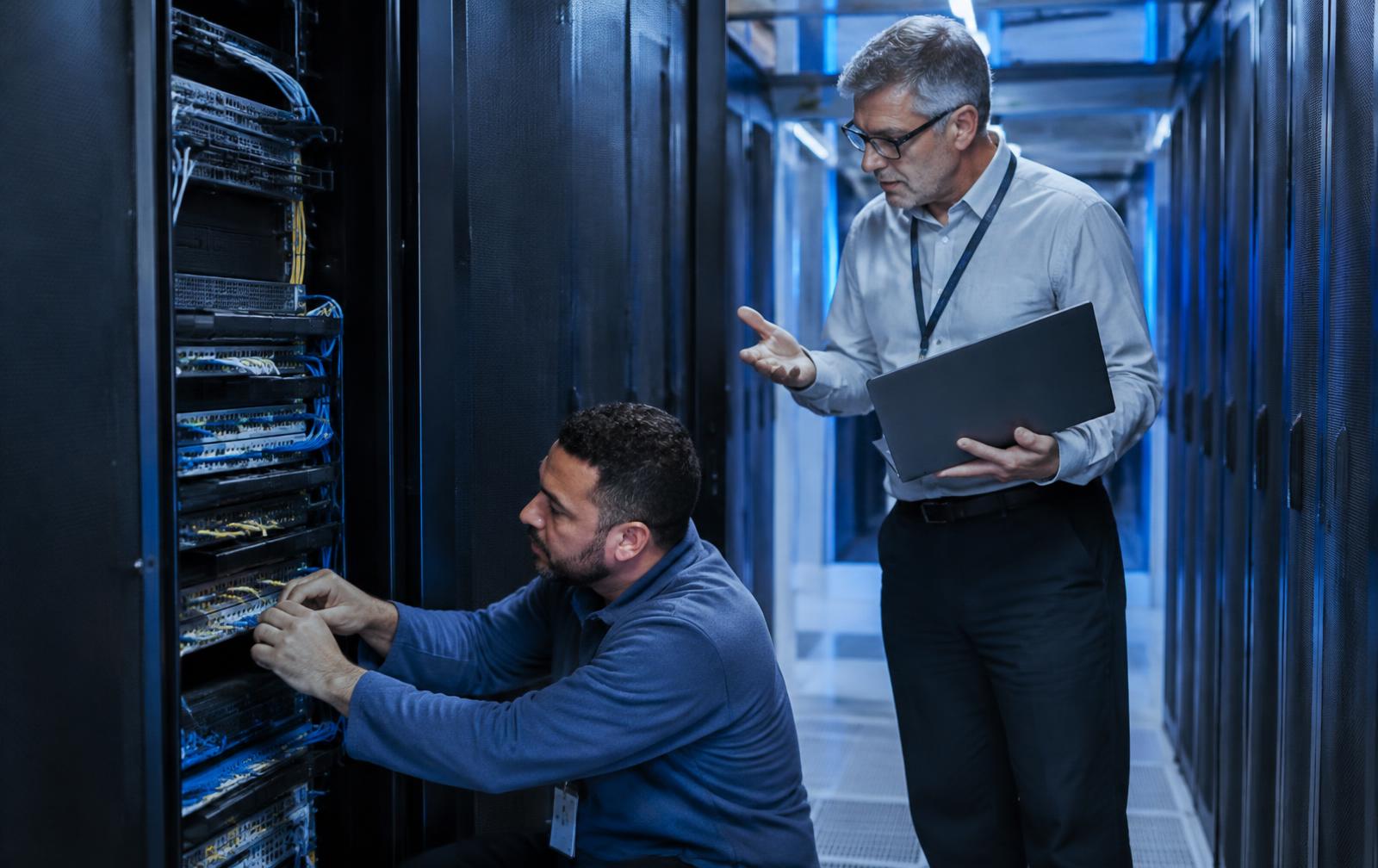 IT infrastructure specialist connecting network cables at a server rack while an in-house engineer with a laptop discusses the work